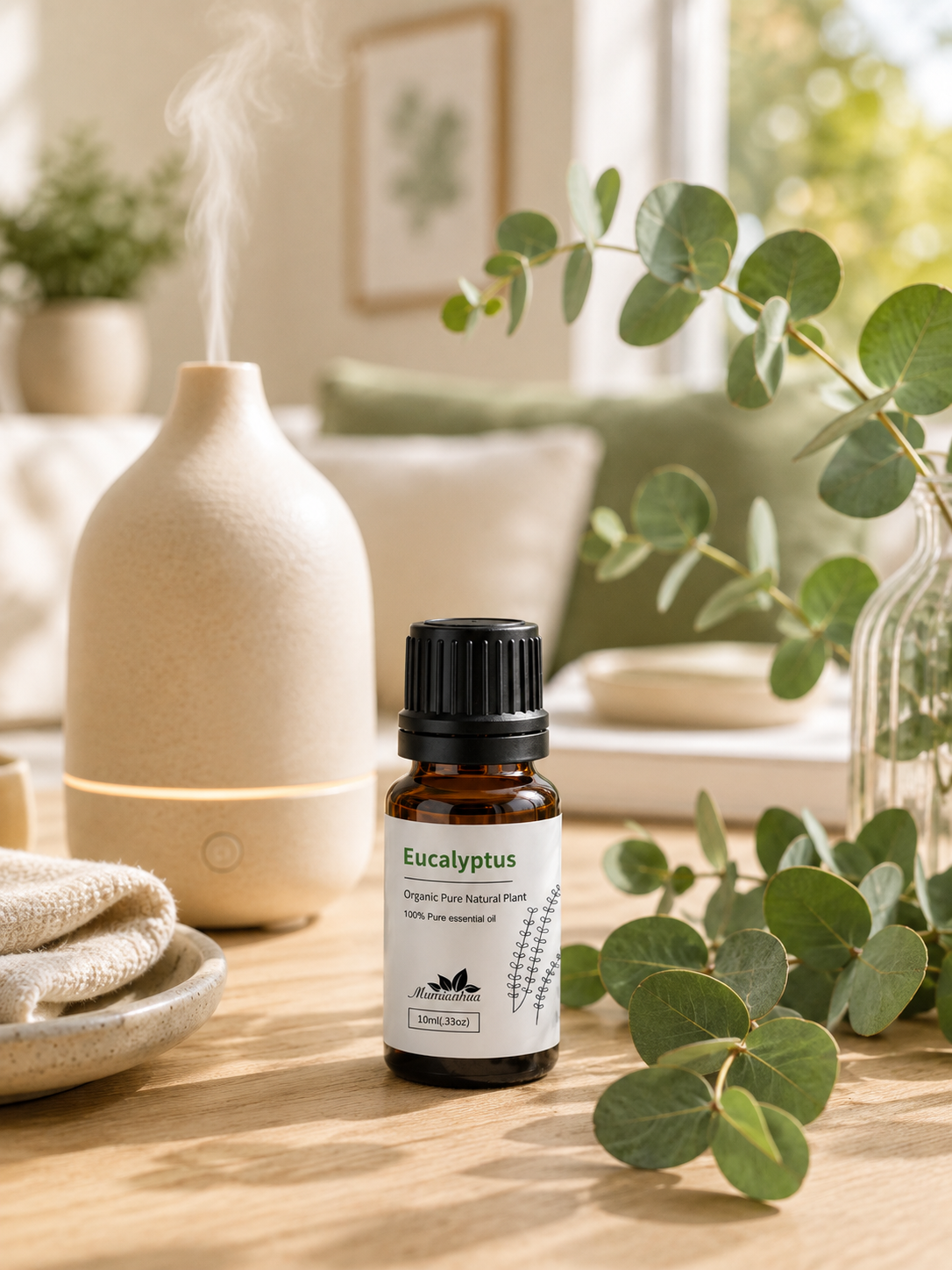 A Eucalyptus amber essential oil bottle with black cap and white label beside a cream ceramic diffuser with visible mist, fresh eucalyptus foliage and soft daylight in a bright airy home